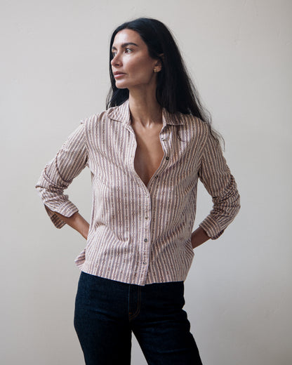 A woman modeling a long-sleeve light pink button-up shirt with vertical stripes and patterns, paired with dark jeans, against a plain light beige background.