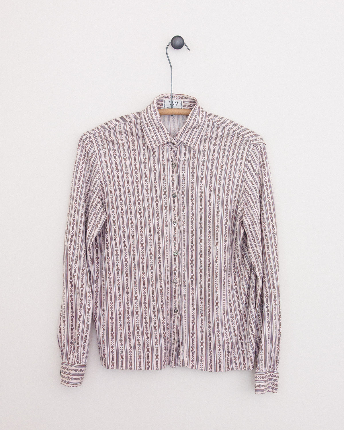 A long-sleeve button-up shirt with horse bit print in vertical stripes in muted pink, beige, white, and brown tones, hanging on a wooden hanger against a plain white wall.