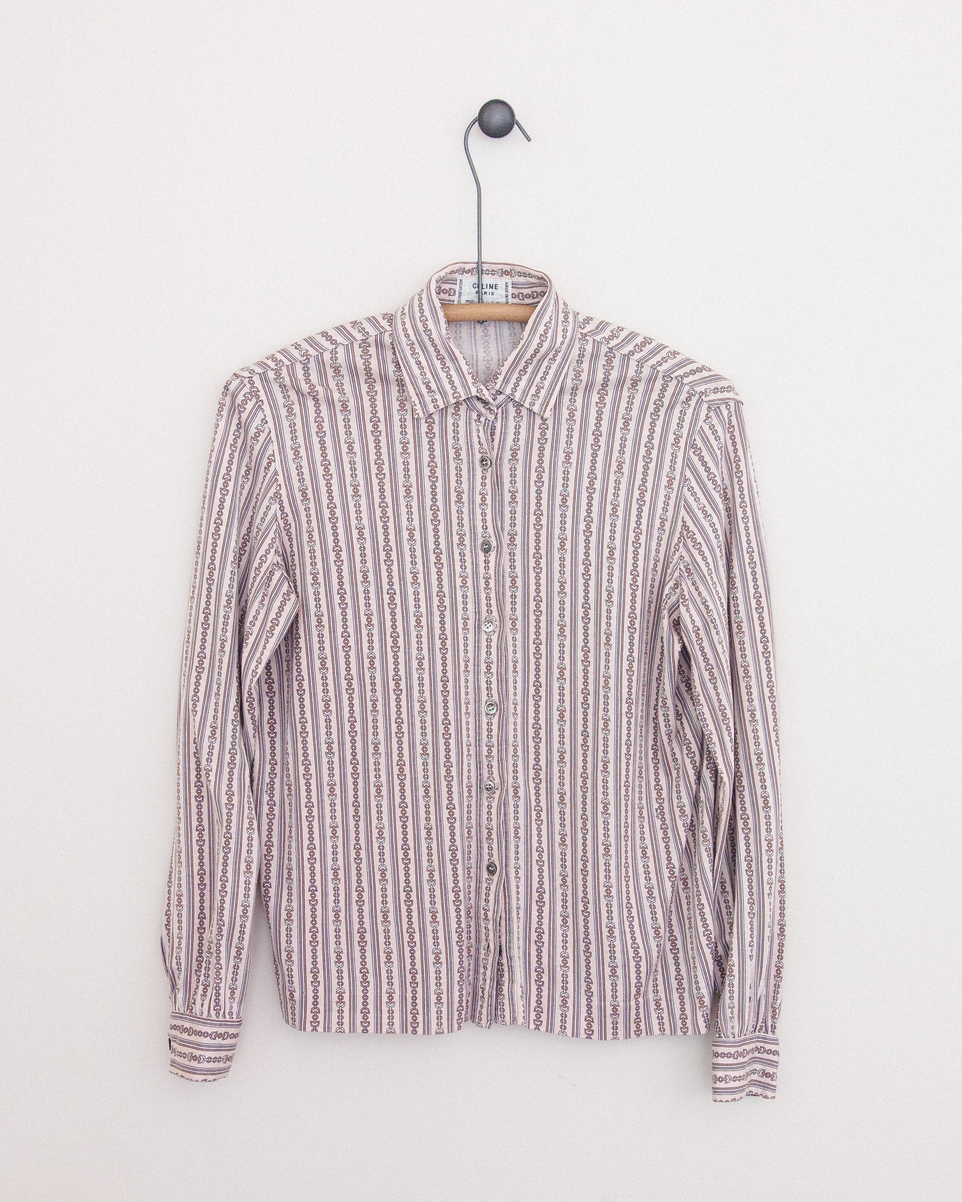 A long-sleeve button-up shirt with horse bit print in vertical stripes in muted pink, beige, white, and brown tones, hanging on a wooden hanger against a plain white wall.