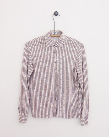 A long-sleeve button-up shirt with horse bit print in vertical stripes in muted pink, beige, white, and brown tones, hanging on a wooden hanger against a plain white wall.