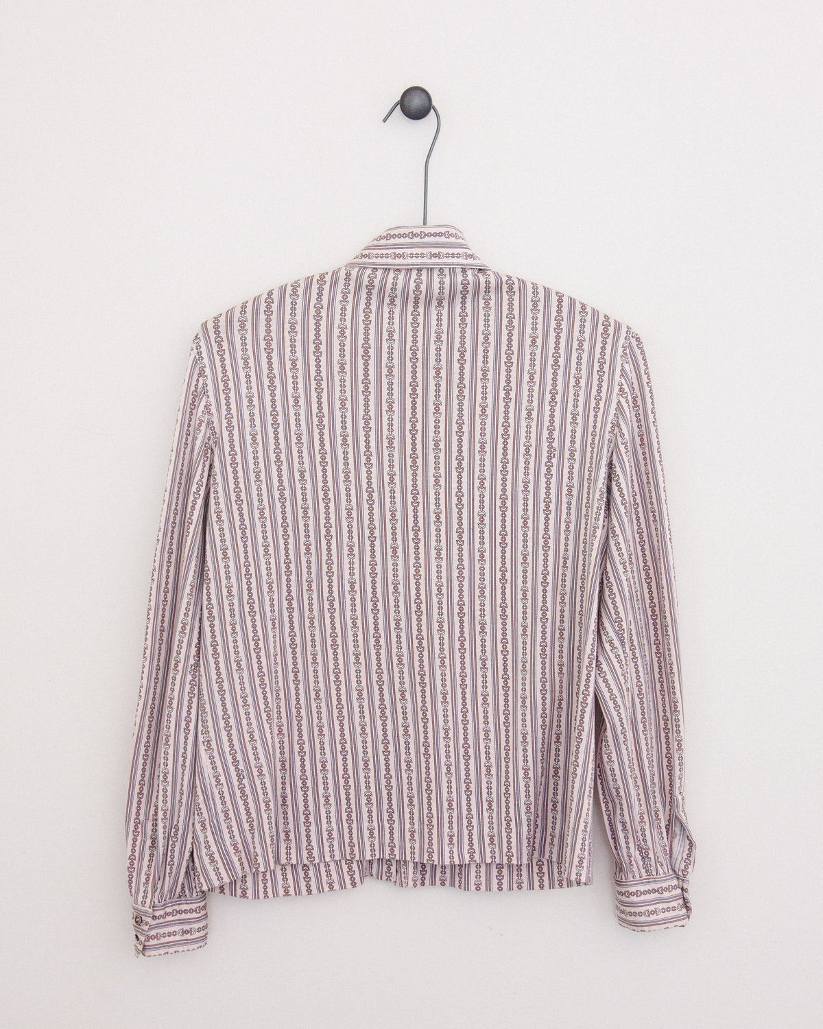A long-sleeve button-up shirt with horse bit pattern in vertical stripes in dusty pink and brown and white, hanging on a black metal hanger against a plain white wall.