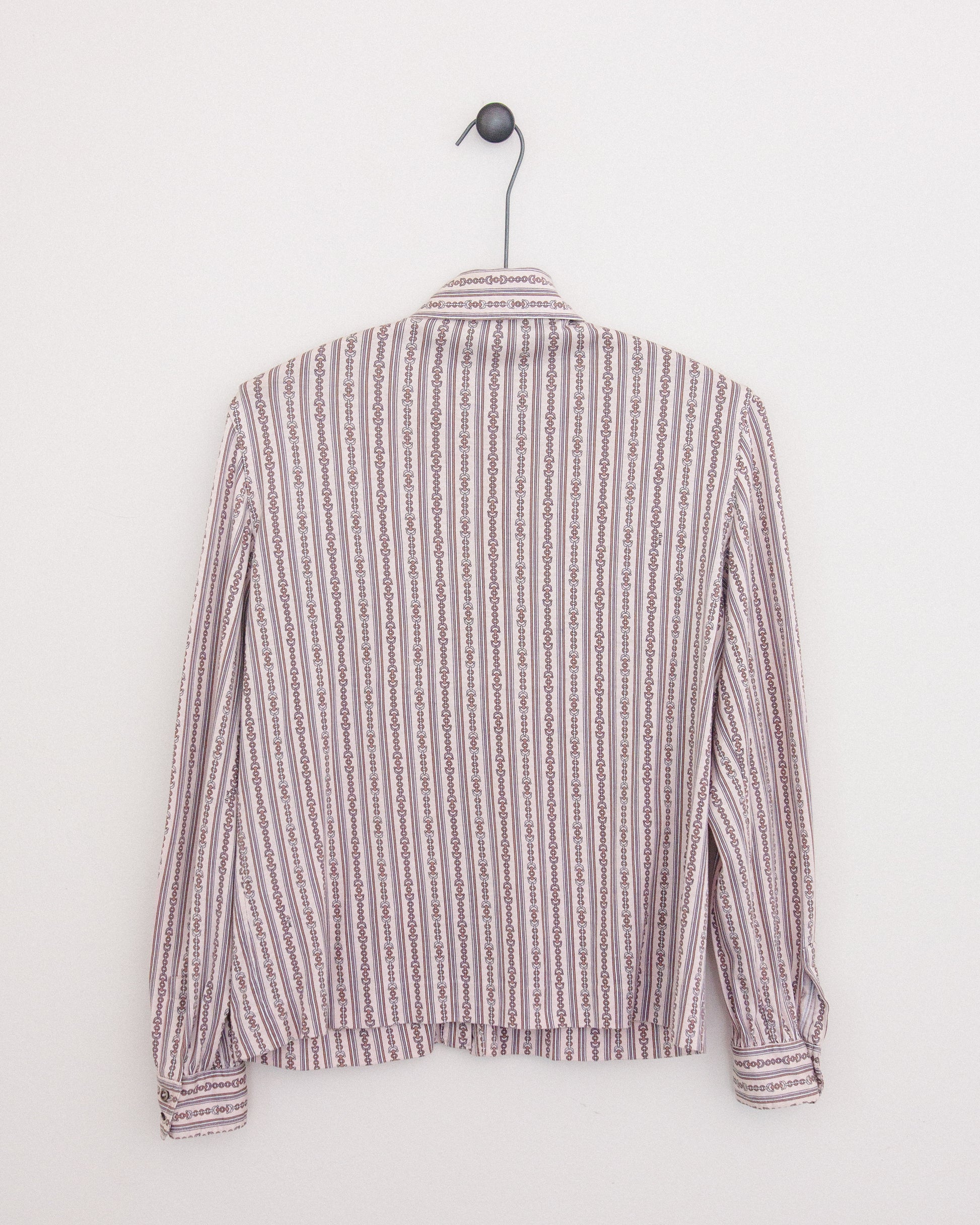 A long-sleeve button-up shirt with horse bit pattern in vertical stripes in dusty pink and brown and white, hanging on a black metal hanger against a plain white wall.