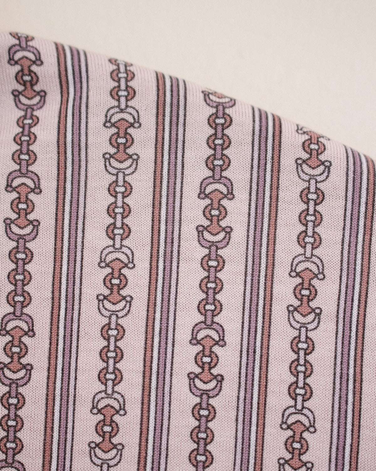 A close-up of a patterned fabric featuring vertical stripes and repeating horse bit chain motifs in muted pink, brown, and white on a light beige background.