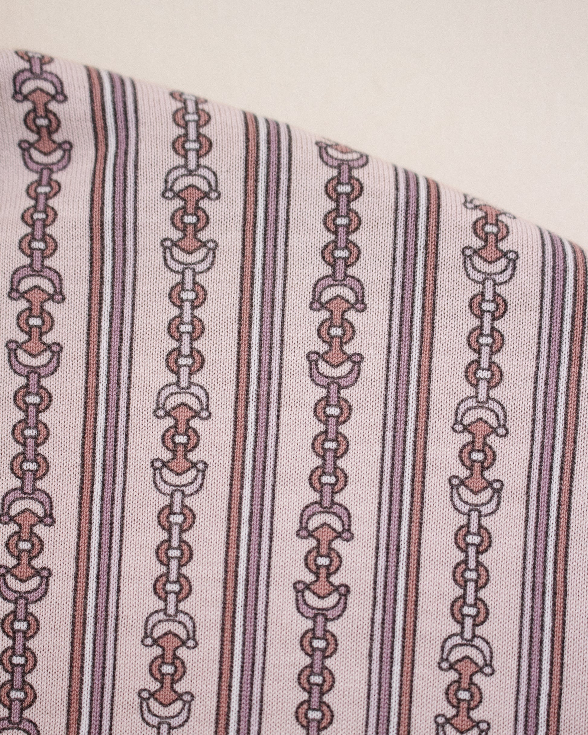 A close-up of a patterned fabric featuring vertical stripes and repeating horse bit chain motifs in muted pink, brown, and white on a light beige background.