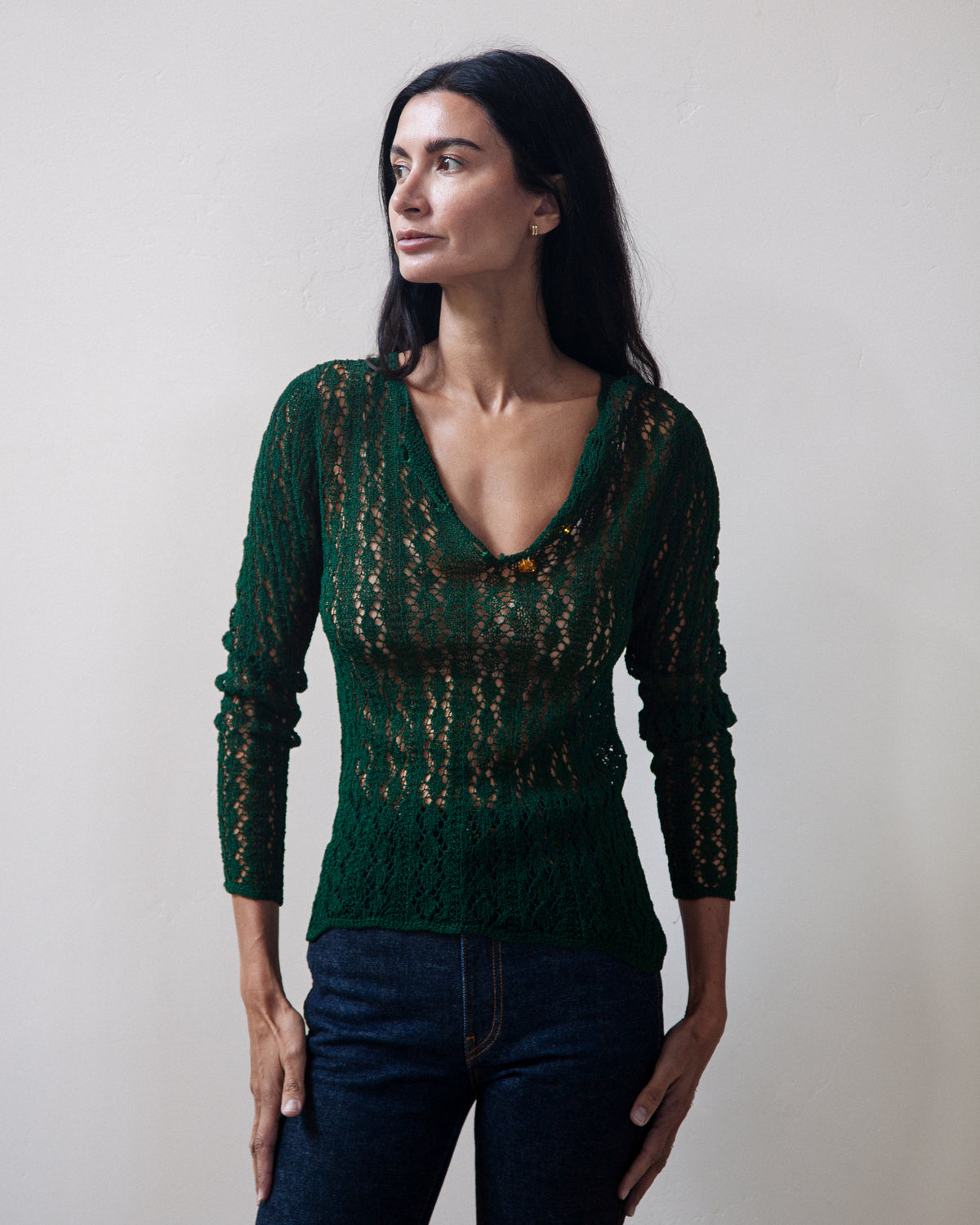 A woman modeling a dark green long-sleeve knit sweater with an open lace pattern, paired with dark blue jeans, against a plain off-white wall.