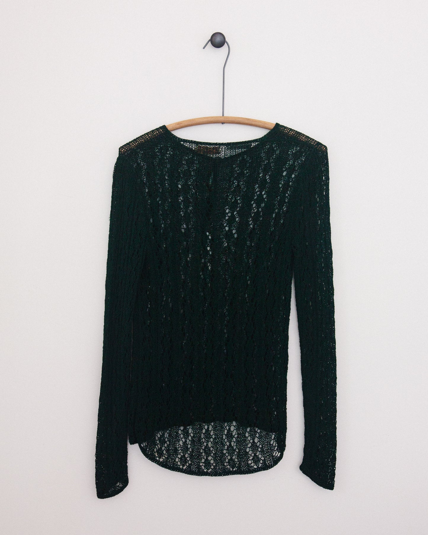 A dark green open-knit long-sleeve top hanging on a wooden hanger against a plain white wall.