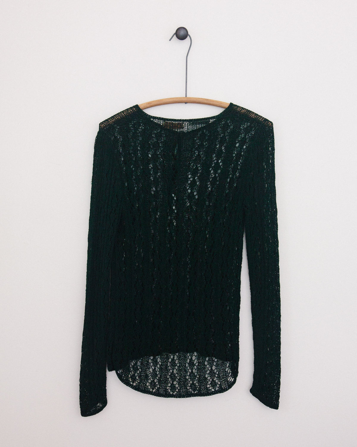 A dark green open-knit long-sleeve top hanging on a wooden hanger against a plain white wall.