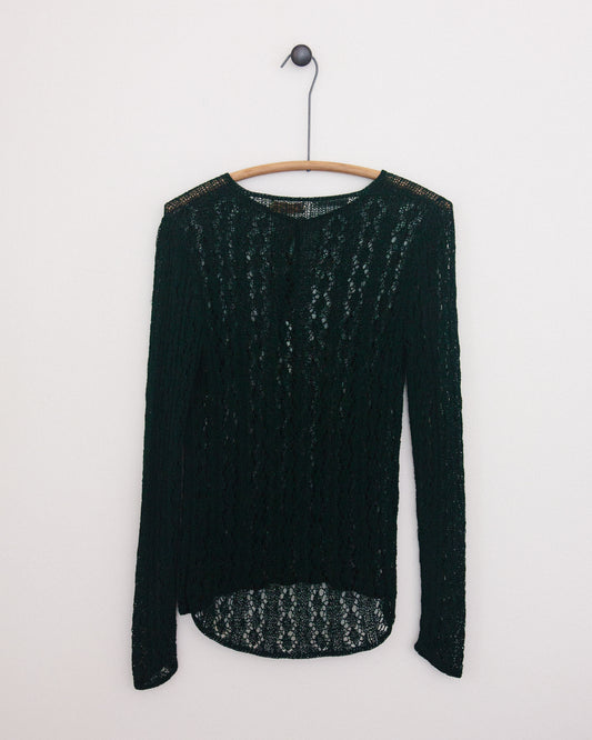 A dark green open-knit long-sleeve top hanging on a wooden hanger against a plain white wall.
