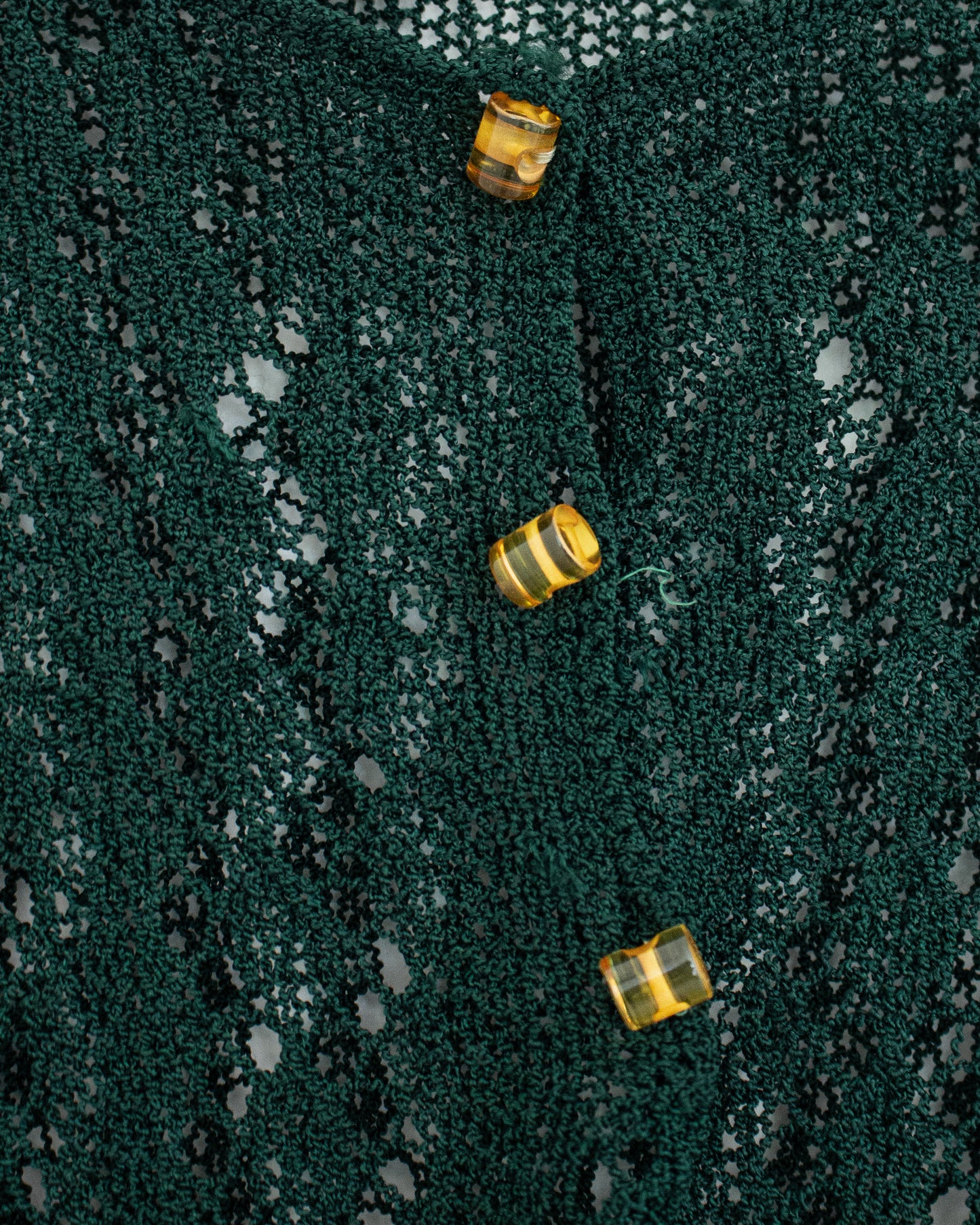Close-up of a dark green open-knit fabric with three small translucent amber-yellow buttons arranged vertically.