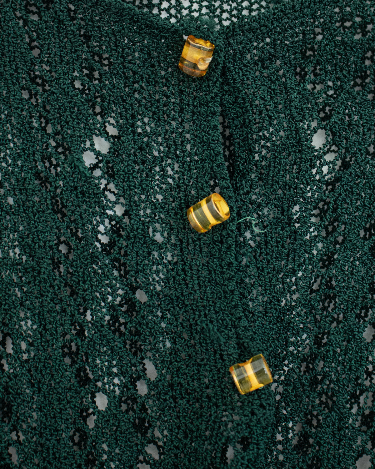 Close-up of a dark green open-knit fabric with three small translucent amber-yellow buttons arranged vertically.