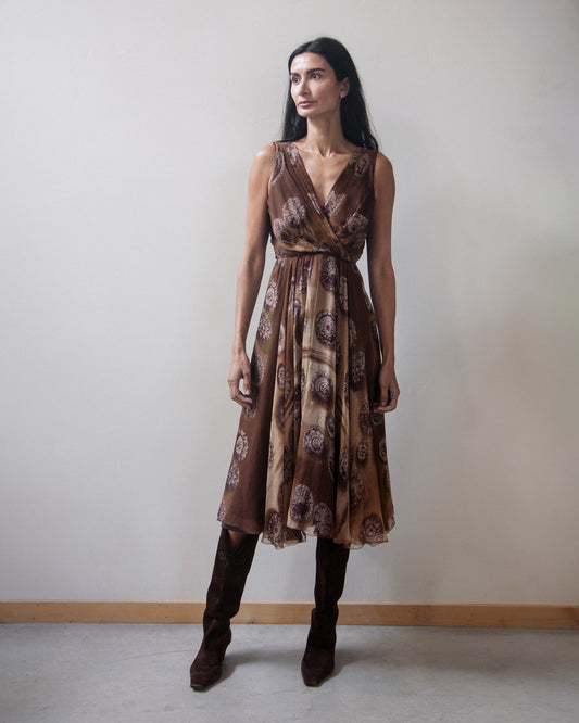 A woman modeling a sleeveless brown wrap-style midi dress with a circular paisley-like print, paired with dark brown knee-high boots, standing against a plain light gray wall.