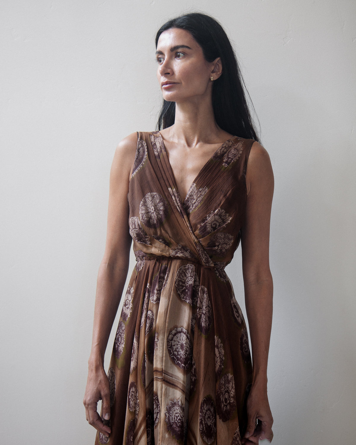 A woman wearing a brown sleeveless wrap-style dress with a brown and beige circular pattern, standing against a plain light gray wall.