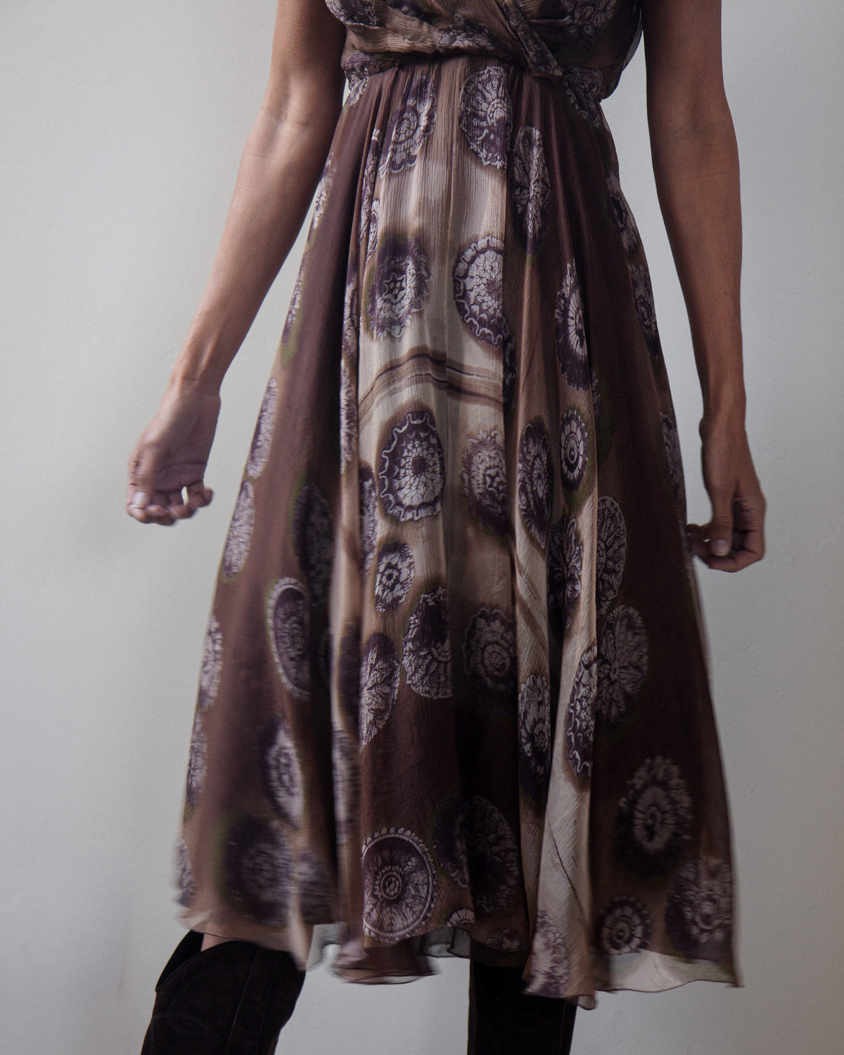 A woman wearing a brown and beige patterned midi dress with a wrap-style bodice and flowing skirt, shown from the waist down against a plain light gray background.