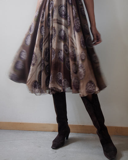 A person wearing a brown and beige patterned dress with dark brown knee-high boots, standing against a plain off-white wall.