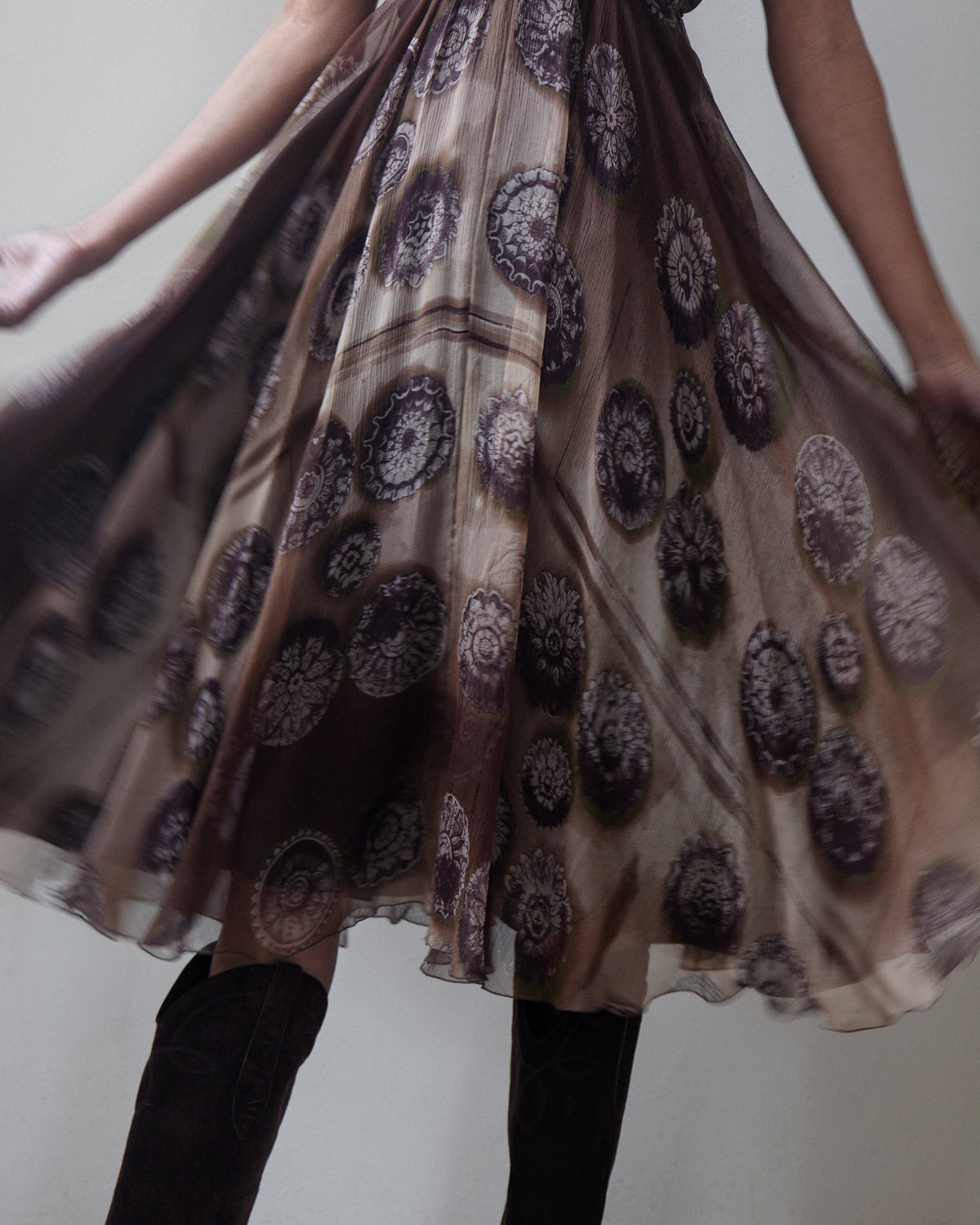 A sheer brown tulle skirt with a dark brown and black circular mandala pattern, worn by a model and flared out. The model is also wearing brown boots, set against a plain light gray background.