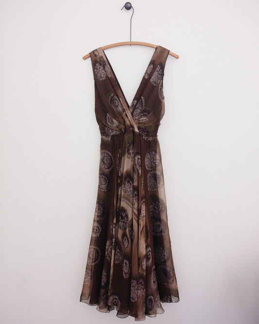 A brown sleeveless dress with a paisley pattern, hanging on a wooden hanger against a plain white wall.