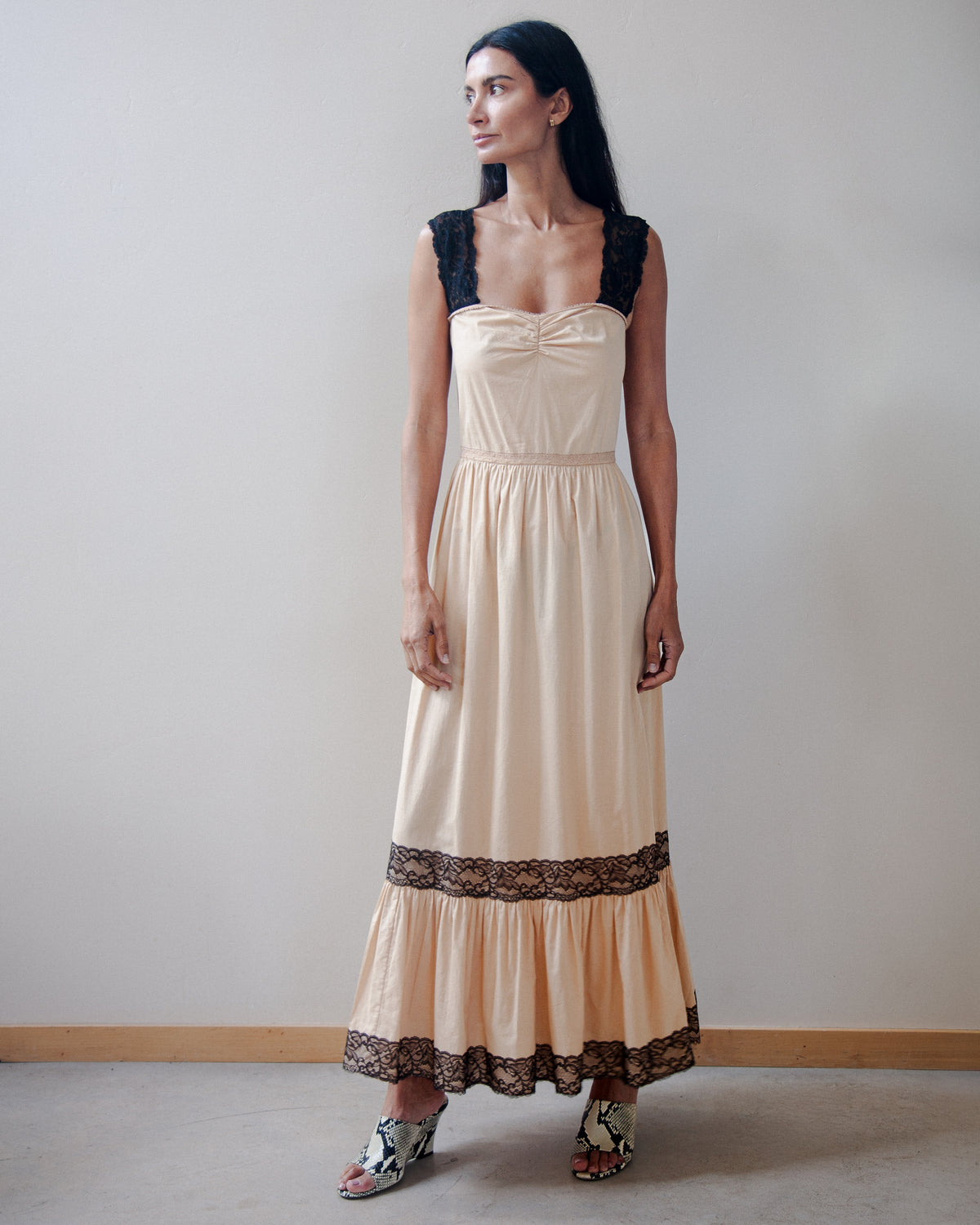 A woman modeling a long beige maxi dress with black lace trim on the bodice and hem, standing against a plain light gray wall.