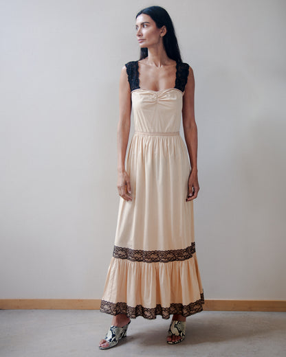 A woman modeling a long beige maxi dress with black lace trim on the bodice and hem, standing against a plain light gray wall.