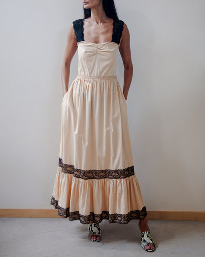 A woman modeling a sleeveless maxi dress with a beige/cream-colored body, black lace shoulder straps, and a matching black lace band near the ruffled hem, standing against a plain light gray wall.