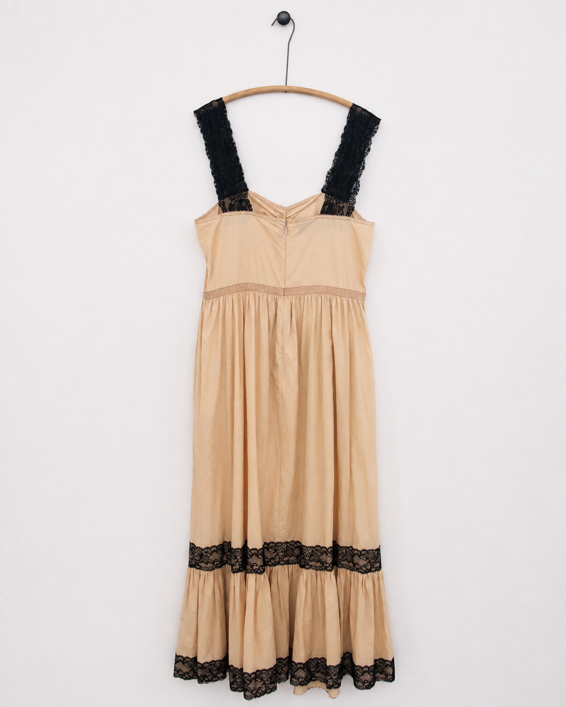 A beige tiered dress with black lace trim on the straps and hem, hanging on a wooden hanger against a plain white wall.