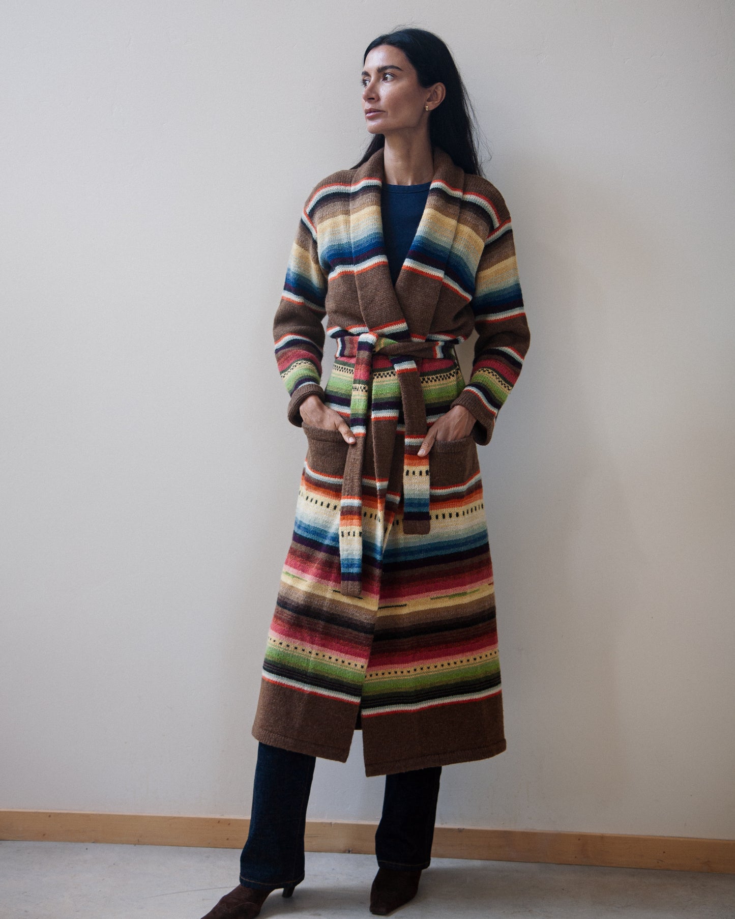 A woman wearing a long, belted striped cardigan with a multicolor serape-inspired pattern, standing against a plain beige wall.
