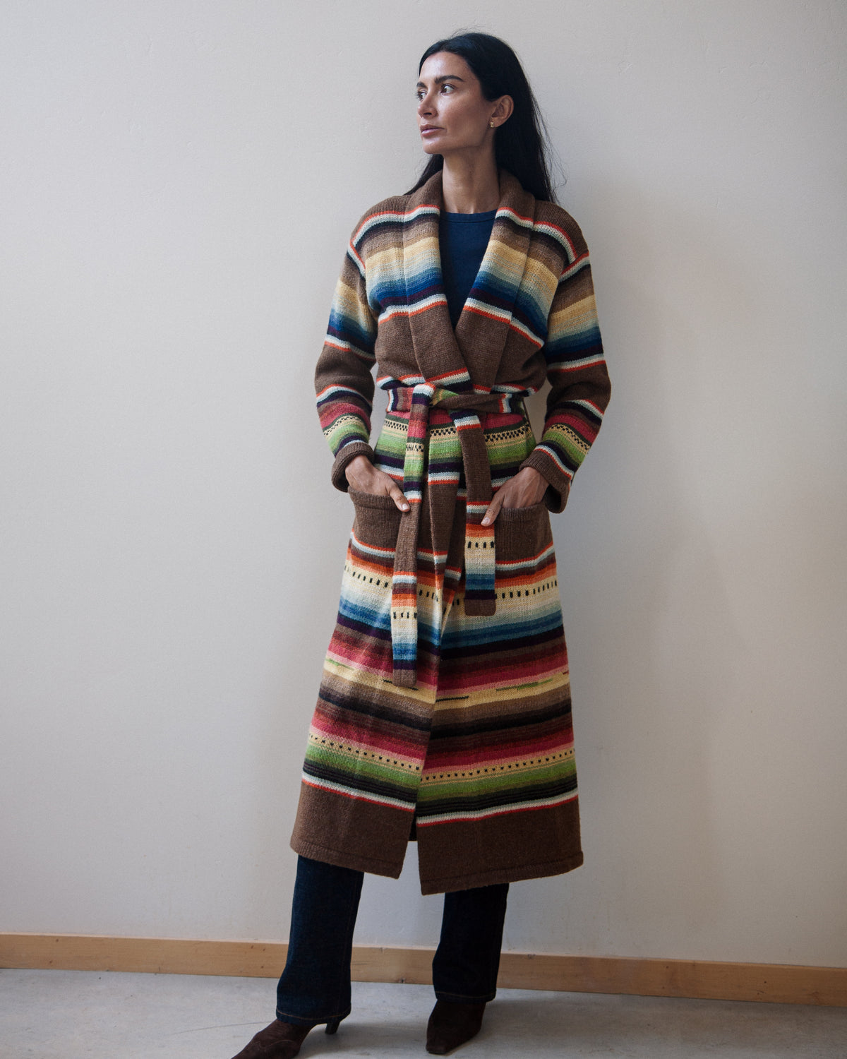 A woman wearing a long, belted striped cardigan with a multicolor serape-inspired pattern, standing against a plain beige wall.