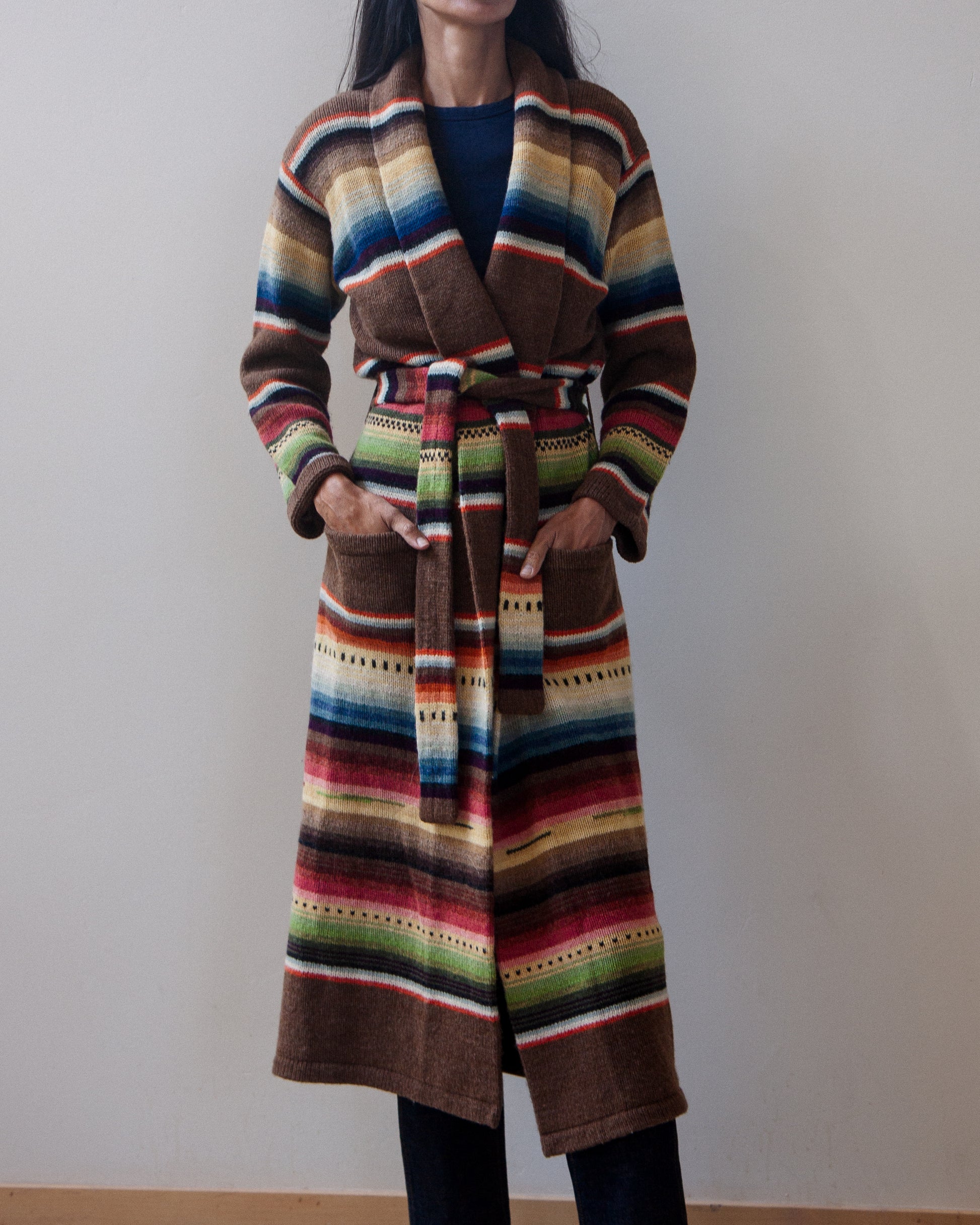 A woman wearing a long, belted striped cardigan with a multicolor serape-inspired pattern, standing against a plain beige wall.