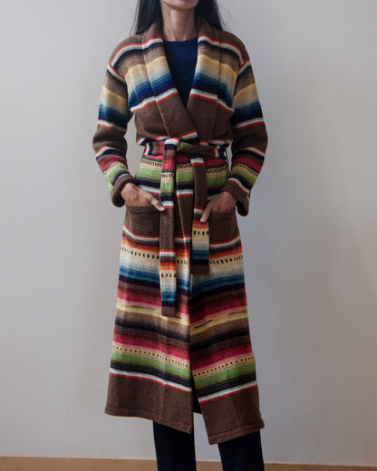 A woman wearing a long, belted striped cardigan with a multicolor serape-inspired pattern, standing against a plain beige wall.