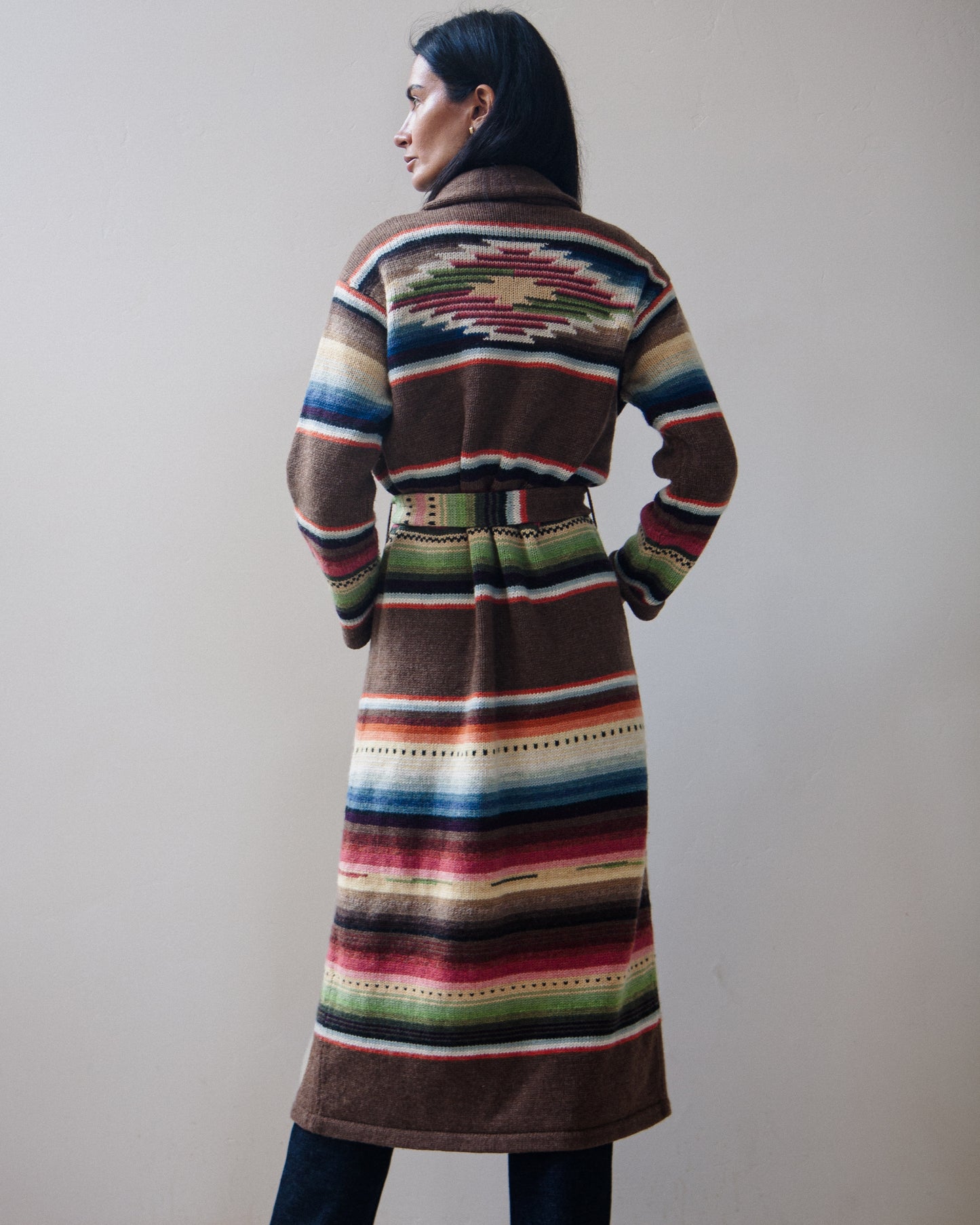 Back view of a woman wearing a long, belted striped cardigan with a multicolor serape-inspired pattern, standing against a plain beige wall.