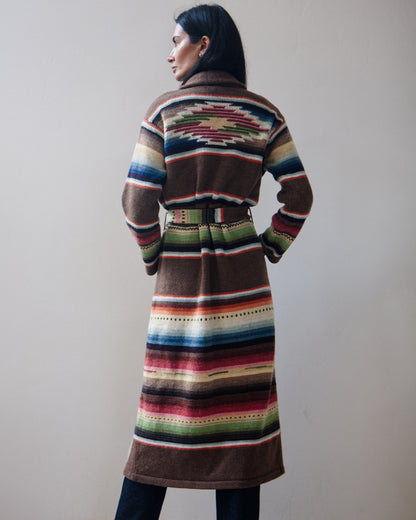 Back view of a woman wearing a long, belted striped cardigan with a multicolor serape-inspired pattern, standing against a plain beige wall.