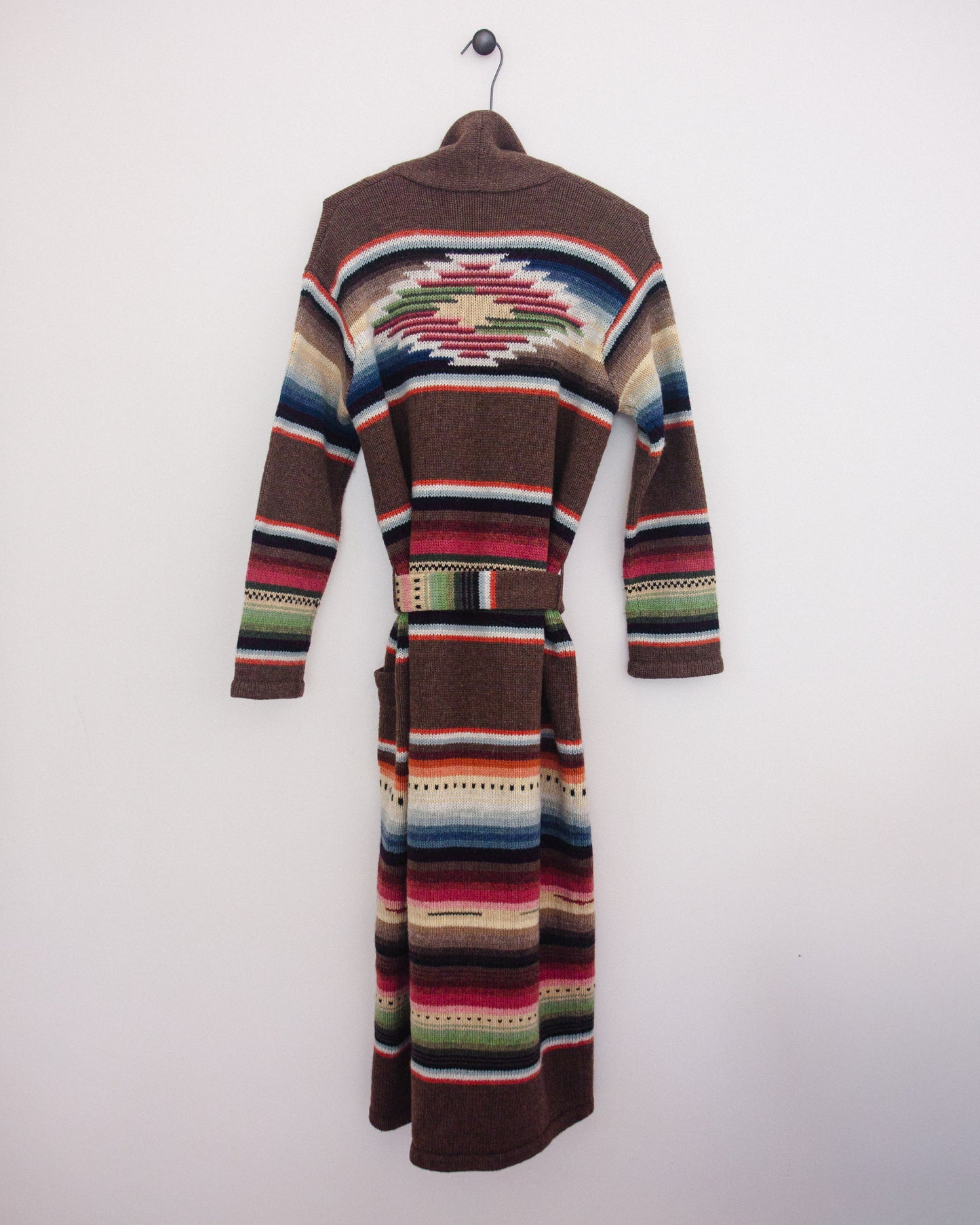 Back of a long brown knit cardigan with Southwestern geometric stripe patterns in red, blue, green, beige, and white, hanging on a black hanger against a plain white wall.