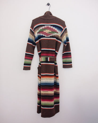 Back of a long brown knit cardigan with Southwestern geometric stripe patterns in red, blue, green, beige, and white, hanging on a black hanger against a plain white wall.