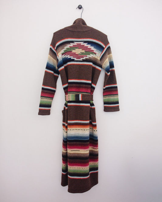 Back of a long brown knit cardigan with Southwestern geometric stripe patterns in red, blue, green, beige, and white, hanging on a black hanger against a plain white wall.