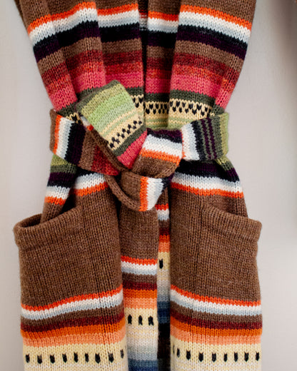 A close-up of a knitted striped garment belt, tied in a bow with two large front pockets. The pattern features horizontal bands of brown, orange, white, black, and green.