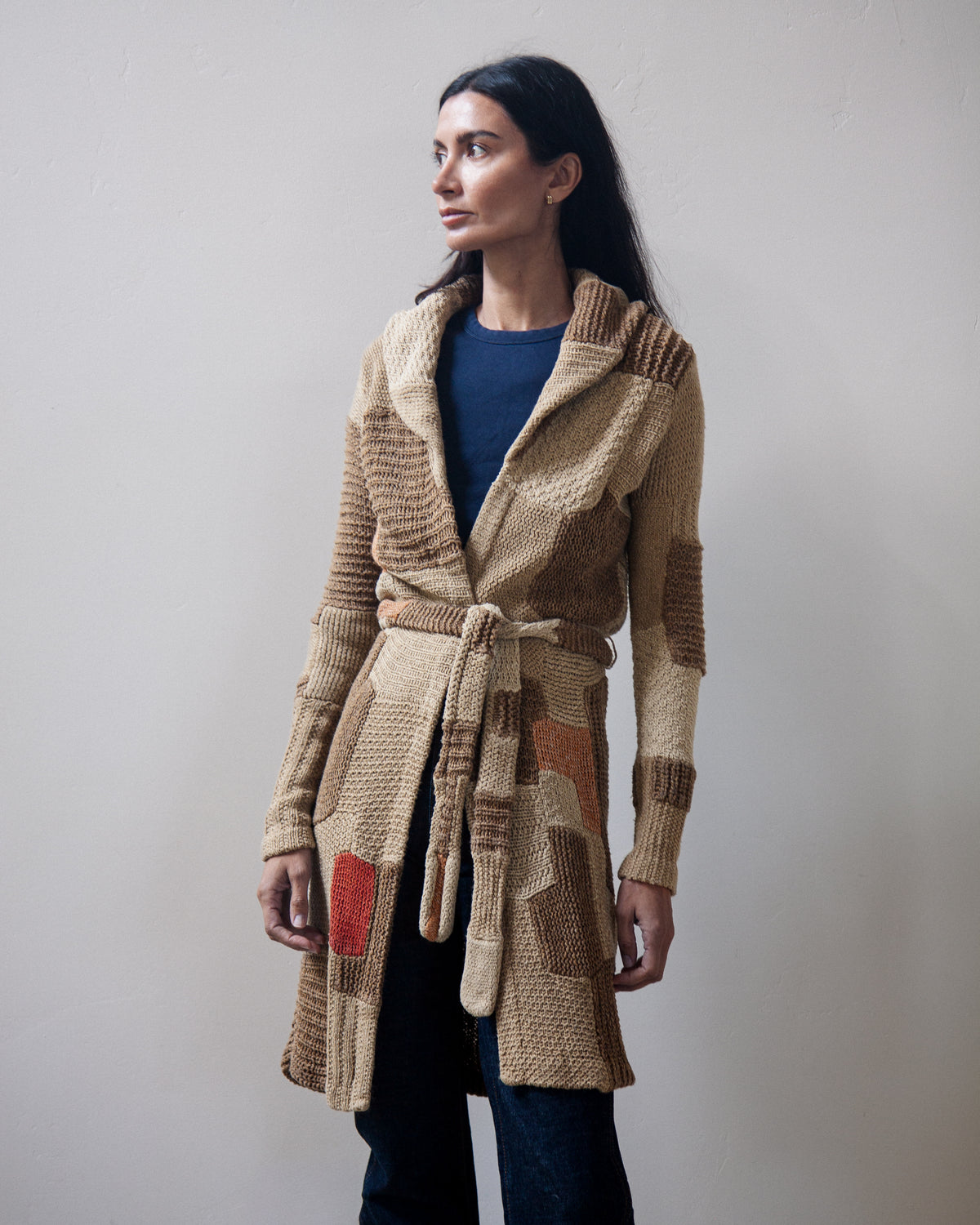A woman wearing a long, chunky knit belted cardigan in beige, brown, and tan tones with a small red accent patch, paired with a navy top and dark pants, standing against a plain light gray wall.