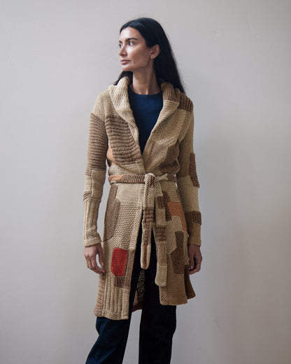 A woman wearing a long, chunky knit belted cardigan in beige, brown, and tan tones with a small red accent patch, paired with a navy top and dark pants, standing against a plain light gray wall.