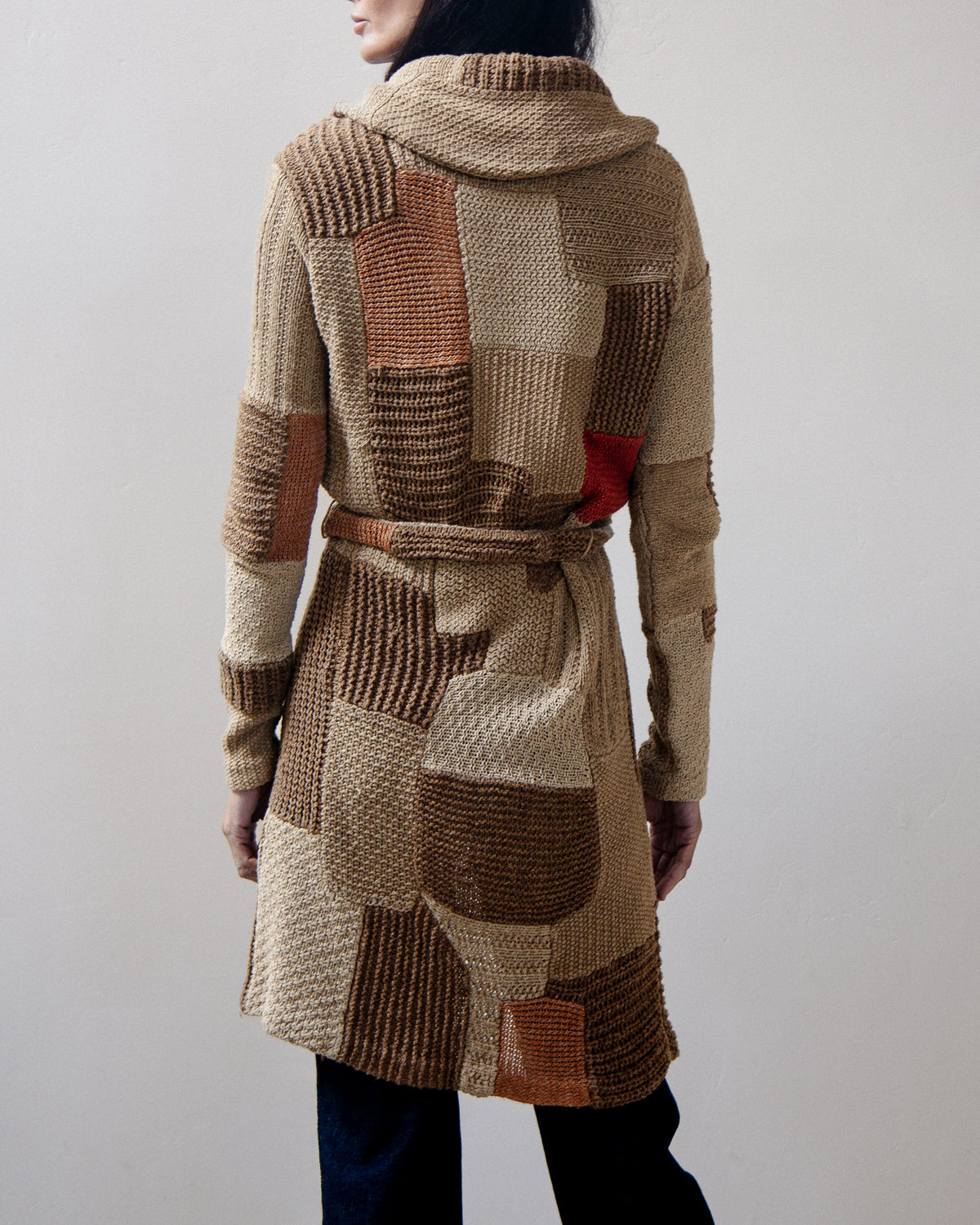 Back view of a person wearing a chunky knit hooded cardigan with patchwork panels in beige, brown, and rust tones, plus a small red accent near the lower back.