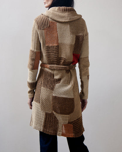 Back view of a person wearing a chunky knit hooded cardigan with patchwork panels in beige, brown, and rust tones, plus a small red accent near the lower back.