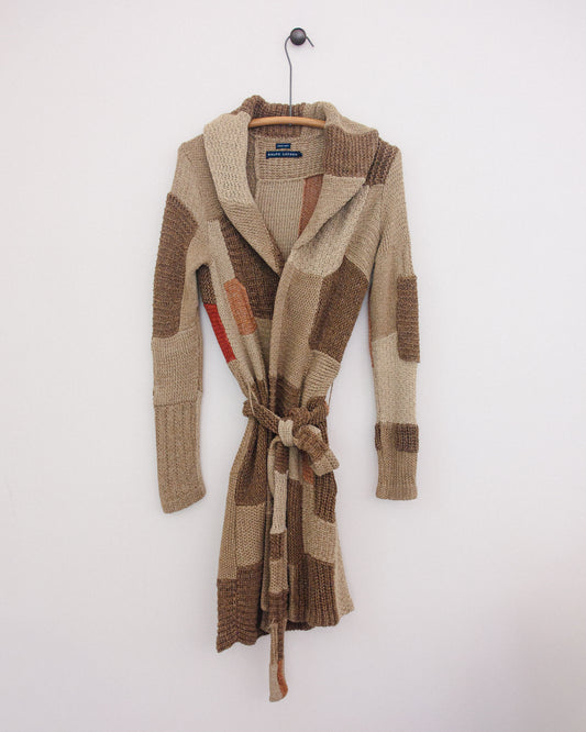 A chunky knit wrap robe with a shawl collar and tie belt, featuring a patchwork design in beige, brown, and tan, hanging on a wooden hanger against a plain white wall.