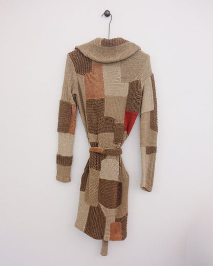 A patchwork knit belted coat with a high mock neck, hanging on a black metal hanger against a plain white wall. The coat features a mix of beige, brown, and terracotta orange textured knit panels, with a small red accent patch on the side.