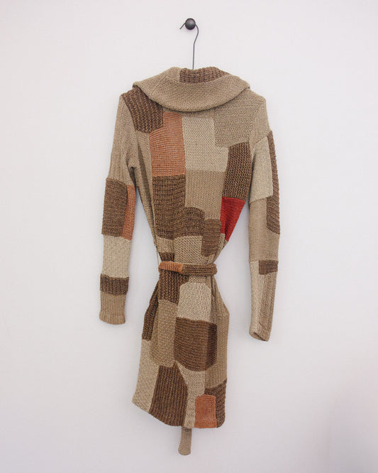 A patchwork knit belted coat with a high mock neck, hanging on a black metal hanger against a plain white wall. The coat features a mix of beige, brown, and terracotta orange textured knit panels, with a small red accent patch on the side.