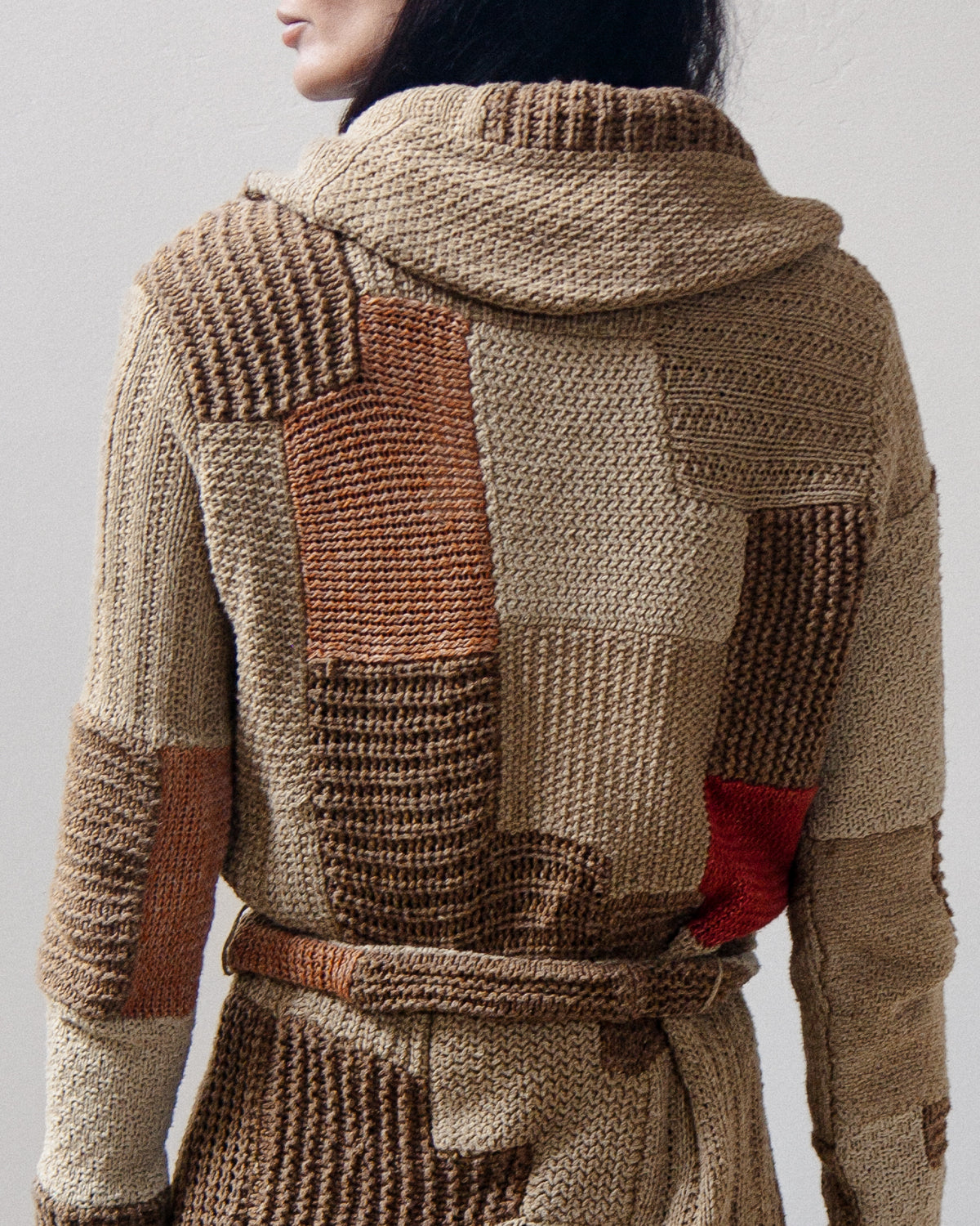 A close-up back view of a person wearing a chunky knit hooded cardigan with patchwork panels in beige, brown, and rust tones, plus a small red accent near the lower back.