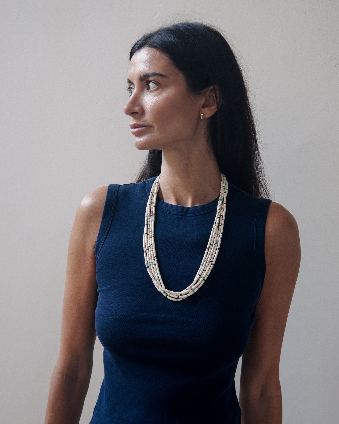 A woman with long dark hair is shown from the chest up, wearing a sleeveless navy top and a layered beaded necklace with cream, white, and light blue beads. She is photographed against a plain, light gray wall.
