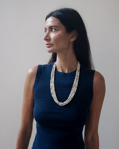 A woman with long dark hair is shown from the chest up, wearing a sleeveless navy top and a layered beaded necklace with cream, white, and light blue beads. She is photographed against a plain, light gray wall.