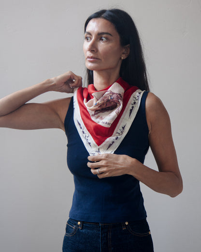 Woman wearing a vintage western-themed scarf featuring a cowboy riding a bucking horse, with the text "LET'ER BUCK" printed below the illustration. The scarf has a bold red border and a white lower section with dark blue ship illustrations.