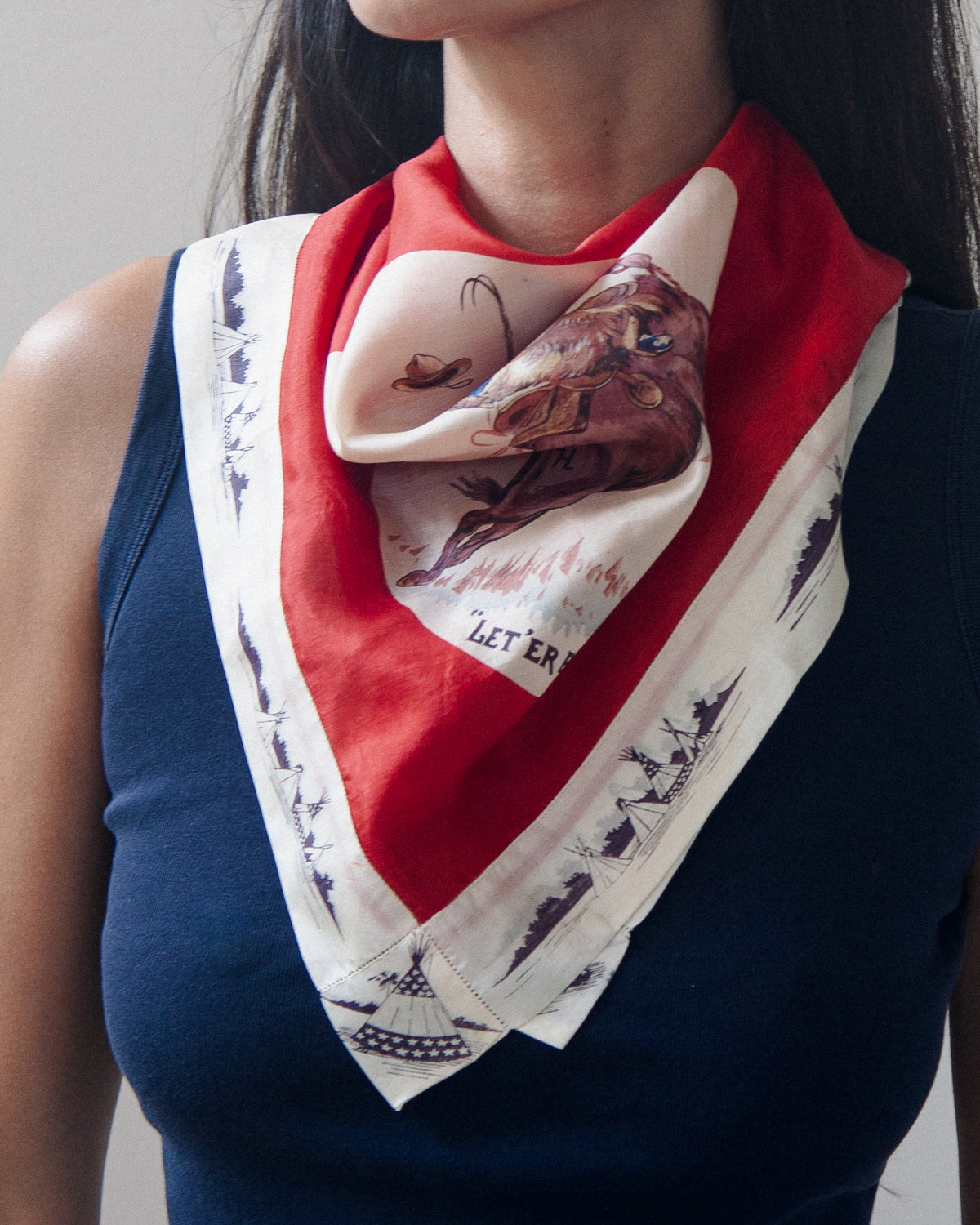 Woman wearing a vintage western-themed scarf featuring a cowboy riding a bucking horse, with the text "LET'ER BUCK" printed below the illustration. The scarf has a bold red border and a white lower section with dark blue ship illustrations.