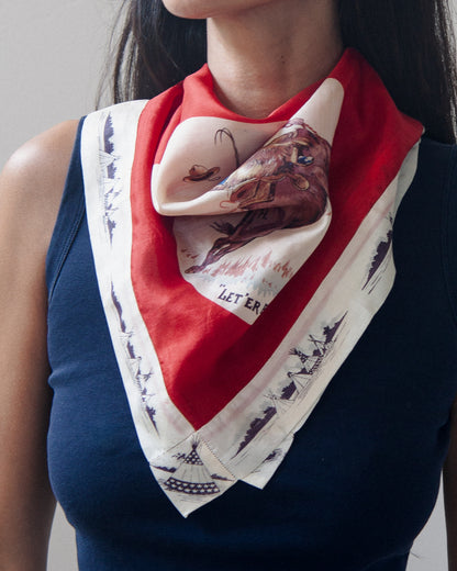 Woman wearing a vintage western-themed scarf featuring a cowboy riding a bucking horse, with the text "LET'ER BUCK" printed below the illustration. The scarf has a bold red border and a white lower section with dark blue ship illustrations.